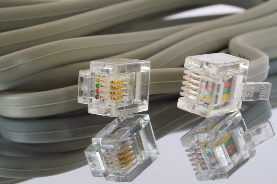 A coiled grey Ethernet cable with four transparent RJ45 connectors resting on a reflective surface. The connectors reveal the small gold-plated pins and multicolored wiring inside. The cable, likely u